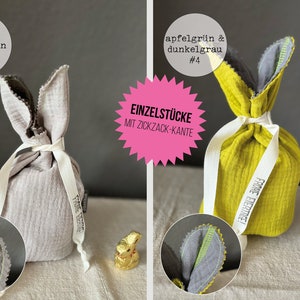 Puede incluir: Dos bolsas de regalo de Pascua de conejo de tela con una cinta blanca. La bolsa de la izquierda es de color gris claro con un borde de zigzag verde. La bolsa de la derecha es de color amarillo brillante con un borde de zigzag verde. Ambas bolsas tienen una etiqueta que dice "Fr&ouml;hliche Eiertage!" que se traduce como "&iexcl;Feliz Pascua!" en ingl&eacute;s.