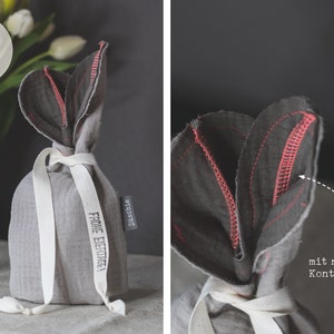 Puede incluir: Una bolsa de regalo de tela gris con una cinta blanca y el texto "Frohe Ostern!" en negro. La bolsa tiene forma de tulip&aacute;n con un detalle de costura rosa.