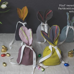 Puede incluir: Cinco bolsas de tela de conejito en diferentes combinaciones de colores. Las bolsas est&aacute;n hechas de una tela suave y texturizada y est&aacute;n atadas con una cinta blanca. Las bolsas est&aacute;n llenas de huevos de chocolate y figuritas de conejito. El texto "F&uuml;nf verschiedene Farbkombinationen erh&auml;ltlich" est&aacute; escrito en alem&aacute;n en el fondo.