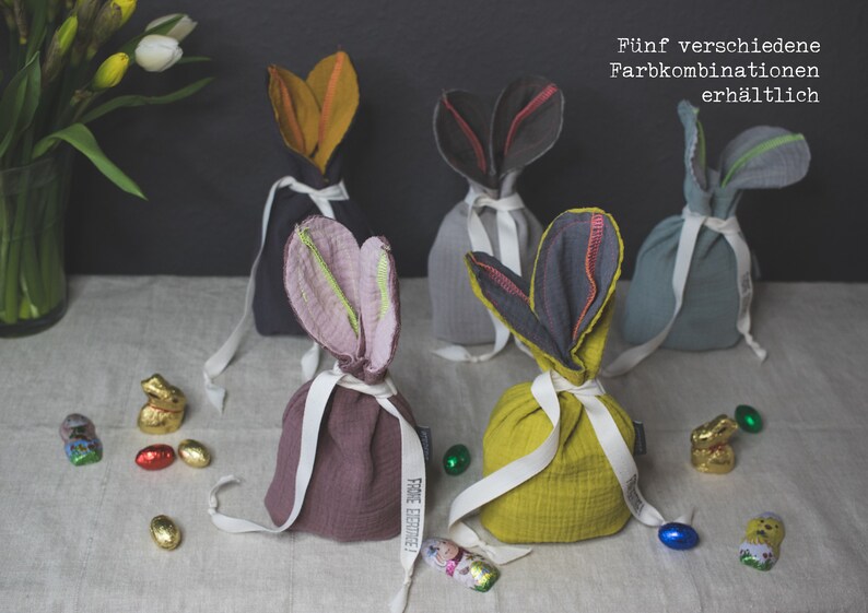 Puede incluir: Cinco bolsas de tela de conejito en diferentes combinaciones de colores. Las bolsas est&aacute;n hechas de una tela suave y texturizada y est&aacute;n atadas con una cinta blanca. Las bolsas est&aacute;n llenas de huevos de chocolate y figuritas de conejito. El texto "F&uuml;nf verschiedene Farbkombinationen erh&auml;ltlich" est&aacute; escrito en alem&aacute;n en el fondo.