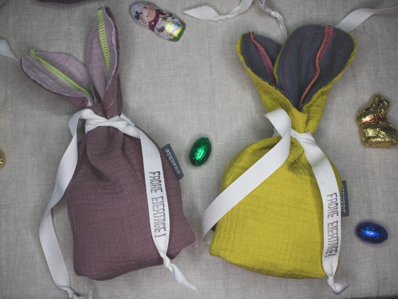Puede incluir: Dos bolsas de regalo de tela con forma de conejo con lazos de cinta blanca. Las bolsas est&aacute;n llenas de huevos de Pascua y tienen el texto "Frohe Ostern!" impreso en ellas.