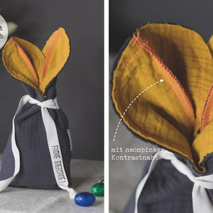 Puede incluir: Una bolsa de regalo de tela gris con un dise&ntilde;o de orejas de conejo de color amarillo mostaza. La bolsa est&aacute; atada con una cinta blanca y tiene el texto "Frohe Ostern!" impreso en el lateral.