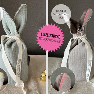 Puede incluir: Dos bolsas de regalo de Pascua de conejo de tela con una cinta blanca y el texto "Frohe Eiertage!" impreso en ellas. La bolsa de la izquierda es azul claro con un borde amarillo en zigzag y la bolsa de la derecha es gris con un borde rosa en zigzag. El texto "Einzelst&uuml;cke mit Zickzack-Kante" est&aacute; impreso en una estrella rosa.