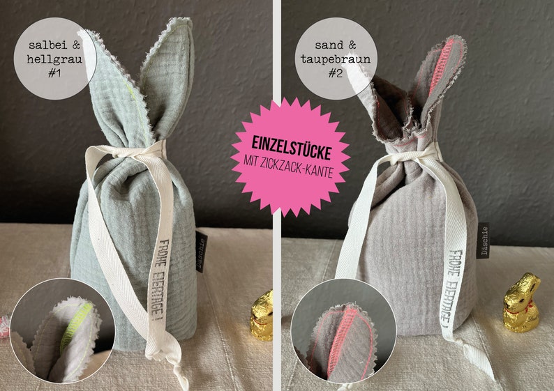 Puede incluir: Dos bolsas de regalo de Pascua de conejo de tela con una cinta blanca y el texto "Frohe Eiertage!" impreso en ellas. La bolsa de la izquierda es azul claro con un borde amarillo en zigzag y la bolsa de la derecha es gris con un borde rosa en zigzag. El texto "Einzelst&uuml;cke mit Zickzack-Kante" est&aacute; impreso en una estrella rosa.