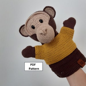 May include: Crocheted monkey puppet wearing a yellow sweater. The puppet has brown fur and a brown nose. The puppet is being held by a hand.