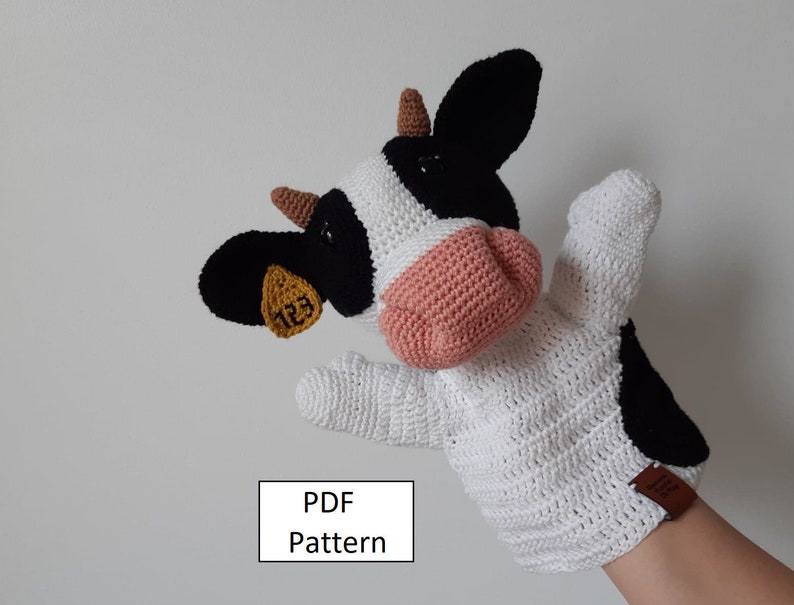 May include: Crocheted cow puppet with black and white spots, a pink nose, and a brown tag with the number 123. The puppet is on a person's hand, with the text "PDF Pattern" below.