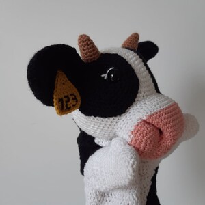 May include: A crocheted stuffed cow toy with black and white fur, brown ears, and a pink nose. The cow has a yellow tag on its ear with the number "123" printed on it.