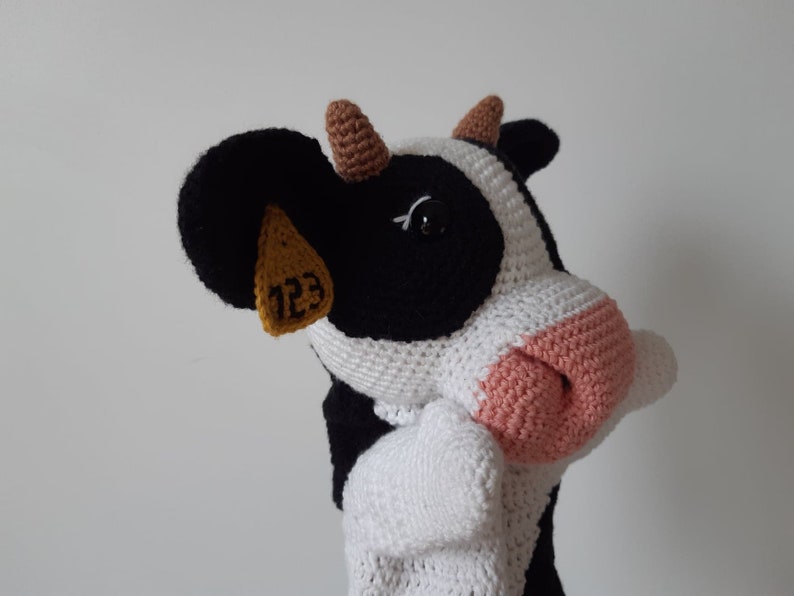 May include: A crocheted stuffed cow toy with black and white fur, brown ears, and a pink nose. The cow has a yellow tag on its ear with the number "123" printed on it.