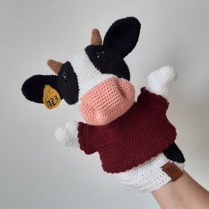 May include: A crocheted cow puppet with black and white spots, a pink nose, and a burgundy sweater. The cow has a yellow tag on its ear with the number "123" printed on it.