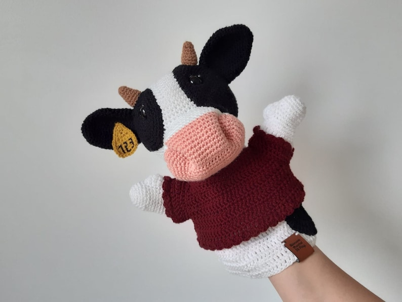 May include: A crocheted cow puppet with black and white spots, a pink nose, and a burgundy sweater. The cow has a yellow tag on its ear with the number "123" printed on it.