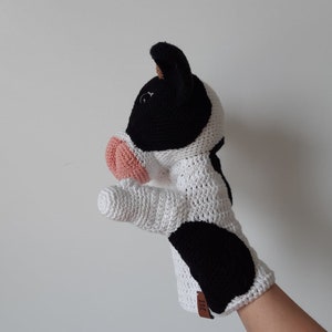 May include: A white and black crocheted cow puppet with a pink nose. The puppet is designed to fit on a hand and has a black and white spotted pattern.