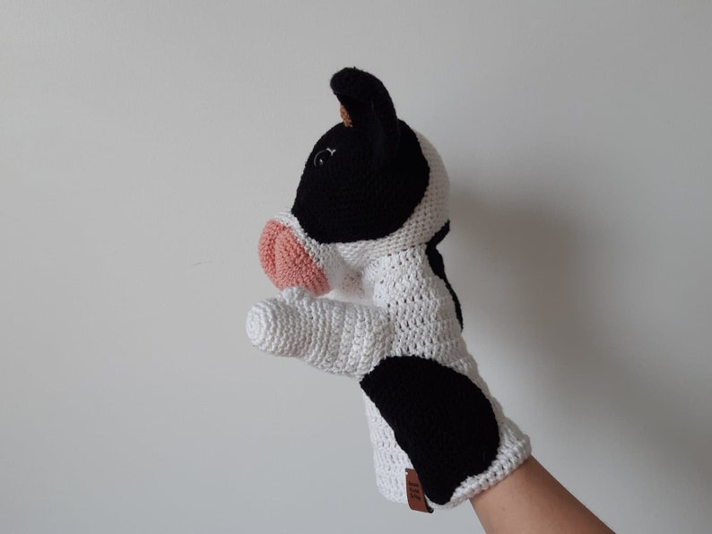 May include: A white and black crocheted cow puppet with a pink nose. The puppet is designed to fit on a hand and has a black and white spotted pattern.