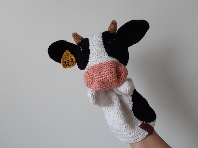 May include: A white and black crocheted cow puppet with a pink nose and a yellow tag with the number 123 on it.