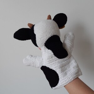 May include: A white and black crocheted cow puppet with brown ears. The puppet is designed to fit on a hand and has a black spot on the side.