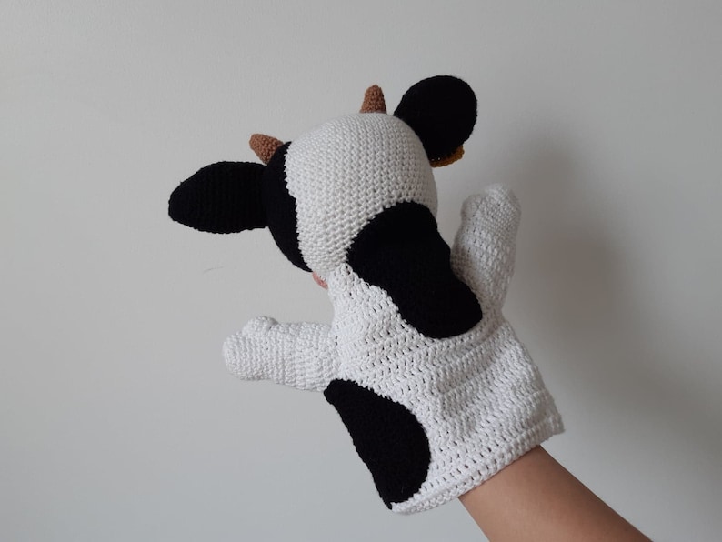 May include: A white and black crocheted cow puppet with brown ears. The puppet is designed to fit on a hand and has a black spot on the side.