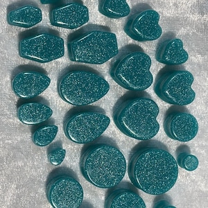 May include: A collection of teal-colored ear plugs in various shapes, including hearts, ovals, and circles. The plugs have a glittery, speckled appearance. The sizes vary, with some small and others larger. They are arranged on a soft, white surface.