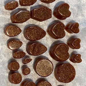 May include: A collection of brown and gold glittery plugs in various shapes, including hexagons, teardrops, hearts, and circles. The plugs are displayed on a white, textured surface. These are likely for ear piercings.