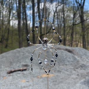 Puede incluir: Adorno de araña con cuentas, suspendido de una cadena dorada. La araña presenta cuentas negras, blancas y transparentes, con una cabeza decorativa y detalles colgantes. El cuerpo es de cuentas transparentes, y las patas de cuentas negras y doradas.