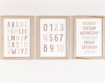 May include: Three framed prints with a white background and light brown frames. The first print features the alphabet in a rainbow of pastel colors. The second print features the numbers 0-10 in a rainbow of pastel colors. The third print features the days of the week in a rainbow of pastel colors.