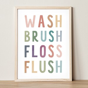 May include: A framed print featuring the words "WASH BRUSH FLOSS FLUSH" in pastel-coloured uppercase letters. The letters are in pink, orange, green, blue, and purple, against a white backdrop. The frame is a light wood colour.