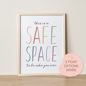 May include: A framed print with the text "This is a safe space to be who you are" in a colorful rainbow font. The print is in a light wood frame and is sitting on a wooden surface.