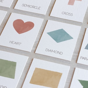 May include: A set of flashcards featuring different geometric shapes, including a heart, diamond, parallelogram, trapezoid, and star. Each card has a watercolor illustration of the shape and the name of the shape written below.