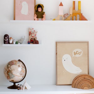 May include: A white shelf with two levels, the top level has a doll wearing a green hooded sweater, a wooden castle, and a framed print of a ghost with the text "Boo!" on a pink background. The bottom level has a globe, a wooden elephant, a wooden reindeer, and a framed print of a ghost with the text "Boo!" on a beige background. The shelf is above three wicker baskets.