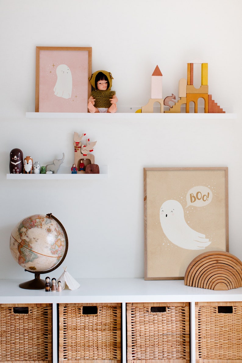 May include: A white shelf with two levels, the top level has a doll wearing a green hooded sweater, a wooden castle, and a framed print of a ghost with the text "Boo!" on a pink background. The bottom level has a globe, a wooden elephant, a wooden reindeer, and a framed print of a ghost with the text "Boo!" on a beige background. The shelf is above three wicker baskets.