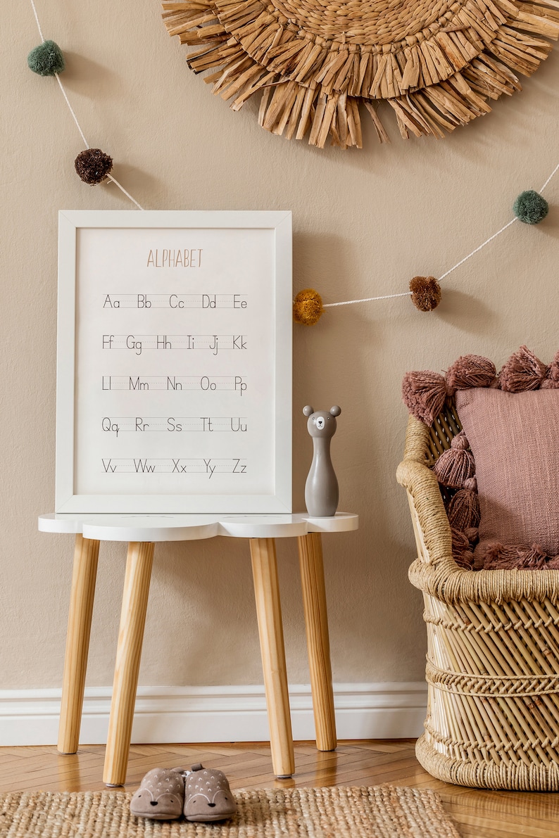 Peut inclure: Un tableau d'alphabet encadr&eacute; blanc avec les lettres de l'alphabet en minuscules, accroch&eacute; &agrave; un mur beige avec une suspension murale en osier au-dessus. Un tabouret blanc avec des pieds en bois se trouve devant le tableau, avec une figurine d'ours en bois gris sur le dessus. Une chaise en osier avec un coussin rose se trouve &agrave; droite du tabouret. Une guirlande de pompons marrons et verts est suspendue en haut du mur.
