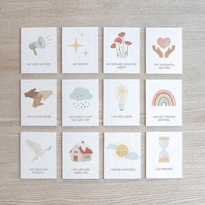 May include: Set of 12 affirmation cards with colorful illustrations and positive messages. The cards feature images of a megaphone, a star, flowers, hands holding a heart, hands shaking, a cloud with raindrops, a lightbulb, a rainbow, a dove, a house, a sun, and an hourglass. The text on the cards includes phrases like "My voice matters", "I am radiant", "My mistakes help me grow", "I am thoughtful and kind", "I am a good friend", "I am perfect just the way I am", "I am intelligent", "I can get through anything", "I am calm and peaceful", "I am safe and cared for", "I choose happiness", and "I am patience".