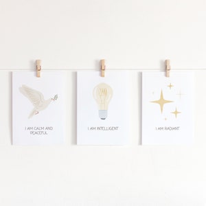 May include: Three white cards with positive affirmations and illustrations. The first card has a white dove with an olive branch in its beak and the text "I am calm and peaceful". The second card has a lightbulb and the text "I am intelligent". The third card has gold stars and the text "I am radiant".