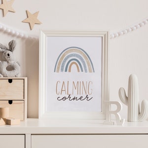 May include: A white framed print with a blue, brown, and white rainbow design and the text "CALMING corner". The print is on a white dresser with a white ceramic cactus and a white letter R.