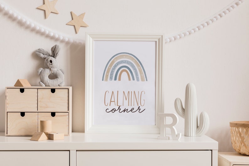 May include: A white framed print with a blue, brown, and white rainbow design and the text "CALMING corner". The print is on a white dresser with a white ceramic cactus and a white letter R.