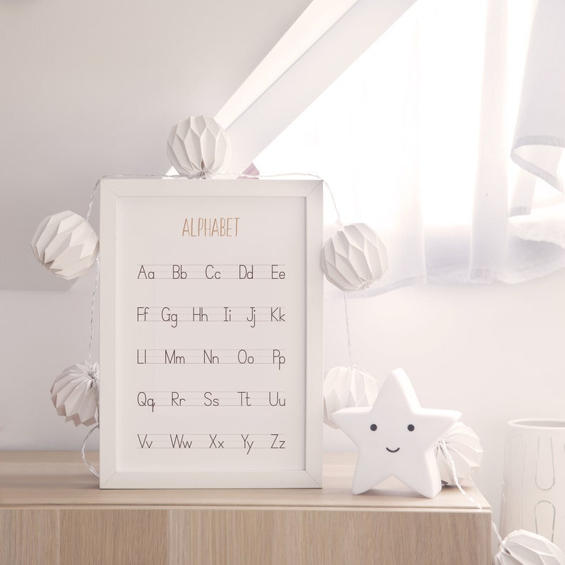 Peut inclure: Une impression encadr&eacute;e blanche avec le mot "ALPHABET" en lettres dor&eacute;es et les lettres de l'alphabet en minuscules, avec deux lettres par ligne. L'impression est entour&eacute;e de lanternes en papier blanc et d'une veilleuse en forme d'&eacute;toile blanche avec un visage souriant.