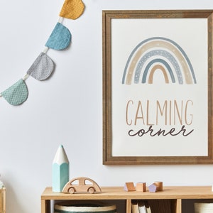 May include: A framed print with a rainbow design and the text "CALMING corner" in a light brown wooden frame. A garland of colorful fabric pennants hangs above the print. A wooden toy car, a blue pencil, and a stuffed animal are on a wooden shelf below the print.