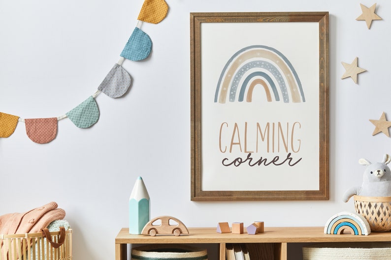 May include: A framed print with a rainbow design and the text "CALMING corner" in a light brown wooden frame. A garland of colorful fabric pennants hangs above the print. A wooden toy car, a blue pencil, and a stuffed animal are on a wooden shelf below the print.
