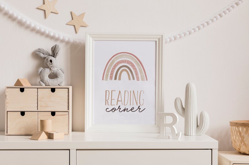 Puede incluir: Una c&oacute;moda blanca con un estampado enmarcado blanco que presenta un arco&iacute;ris con tonos marrones y rosados y el texto "READING corner". Hay un cactus blanco, una letra R de madera y un conejo de juguete de madera en la c&oacute;moda.