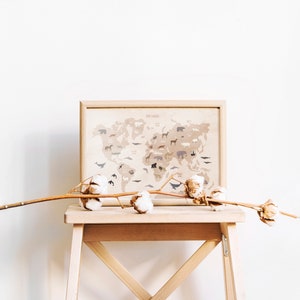 May include: A framed world map with animal illustrations printed on a beige background. The map is displayed on a wooden stool with a dried cotton branch on top.