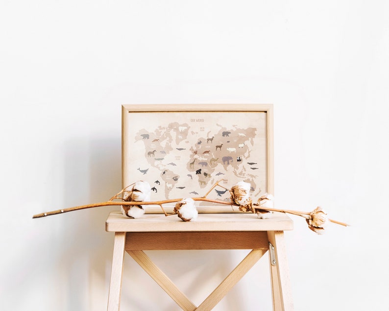 May include: A framed world map with animal illustrations printed on a beige background. The map is displayed on a wooden stool with a dried cotton branch on top.