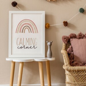 Puede incluir: Una impresi&oacute;n enmarcada en blanco con un arco&iacute;ris y el texto "CALMING corner" sobre una mesa blanca con patas de madera. Una peque&ntilde;a figurita de oso gris est&aacute; sobre la mesa. Una silla de mimbre con un coj&iacute;n rosa est&aacute; en el fondo.
