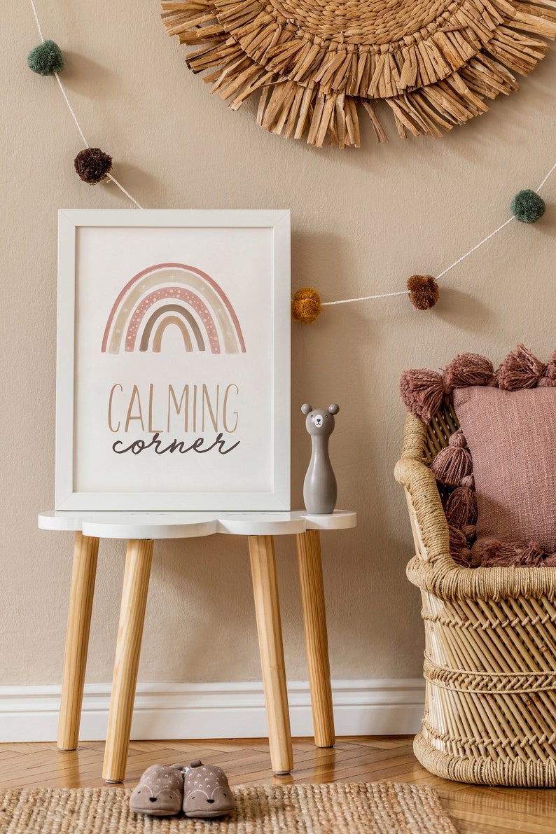 Puede incluir: Una impresi&oacute;n enmarcada en blanco con un arco&iacute;ris y el texto "CALMING corner" sobre una mesa blanca con patas de madera. Una peque&ntilde;a figurita de oso gris est&aacute; sobre la mesa. Una silla de mimbre con un coj&iacute;n rosa est&aacute; en el fondo.