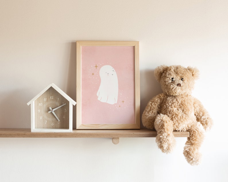 May include: A white wooden house-shaped clock with black hands and a pink and white framed print of a ghost with gold stars on a pink background. A brown teddy bear sits on a wooden shelf.