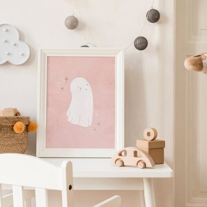May include: A white framed picture with a pink background and a white ghost illustration. A white cloud light with white bulbs, a brown wicker basket with orange pom-poms, and a wooden toy car are also in the image.