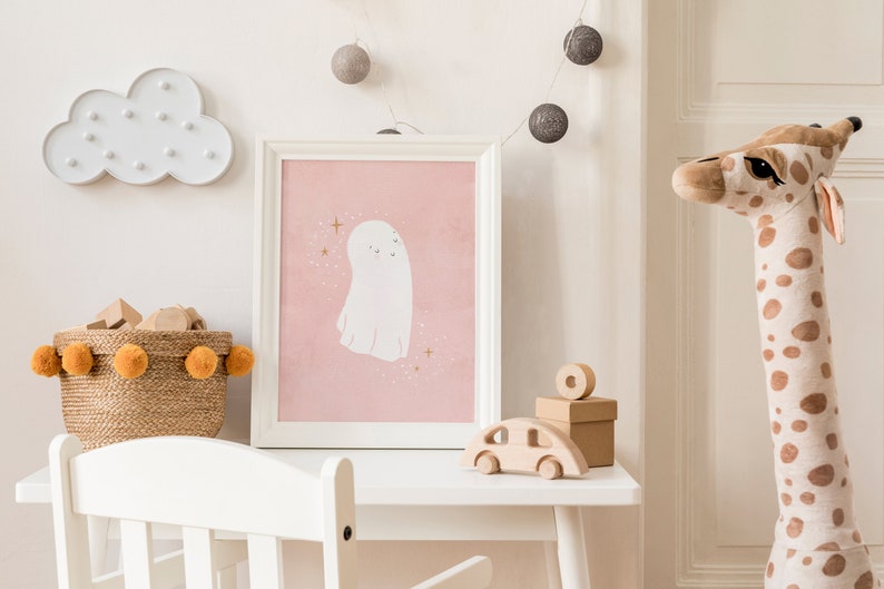 May include: A white framed picture with a pink background and a white ghost illustration. A white cloud light with white bulbs, a brown wicker basket with orange pom-poms, and a wooden toy car are also in the image.
