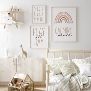 Puede incluir: Una habitaci&oacute;n de ni&ntilde;os con una pared blanca y tres impresiones enmarcadas. Las impresiones presentan un arco&iacute;ris con colores marrones y rosas, las palabras "PLAY all DAY" y los n&uacute;meros del 0 al 10. La habitaci&oacute;n tambi&eacute;n tiene un avi&oacute;n de juguete de madera, una casa de mu&ntilde;ecas de madera, una jirafa de madera y una l&aacute;mpara blanca.