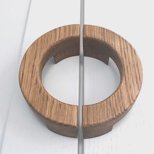 Peut inclure: Une paire de poignées de placard en bois. Chaque poignée est en forme de demi-cercle avec une finition en grain de bois naturel. Les poignées sont montées sur une surface blanche, avec une fine bande métallique entre elles.