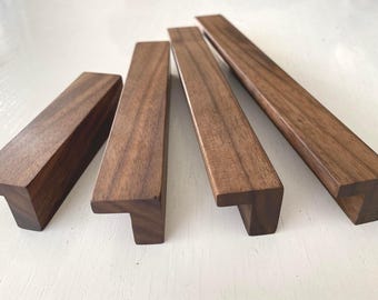 Può includere: Quattro maniglie per mobili in legno con finitura marrone scuro. Le maniglie hanno una forma rettangolare con una maniglia a forma di T. La venatura del legno è visibile. Probabilmente per mobili da cucina o bagno.