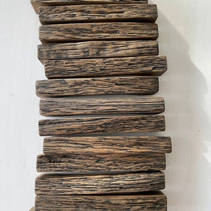 Op de afbeelding: Een stapel van 14 gerecyclede houten stukken, elk met een uniek houtnerfpatroon en een donkerbruine kleur. De houten stukken zijn in een verspringende stapel gelegd, wat een visueel aantrekkelijk display creëert.