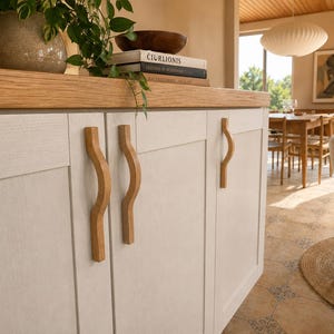 May include: Off-white kitchen cabinetry featuring a light wood grain texture and vertical light oak cabinet pulls with a curved, wave-like design. The cabinets are topped with a thick, natural light oak countertop surface.