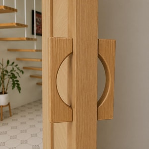 Peut inclure: Une poignée de porte en bois clair présentant une base rectangulaire avec une découpe semi-circulaire formant une prise incurvée. La poignée est montée verticalement sur un panneau de porte en bois clair assorti avec un grain naturel visible.